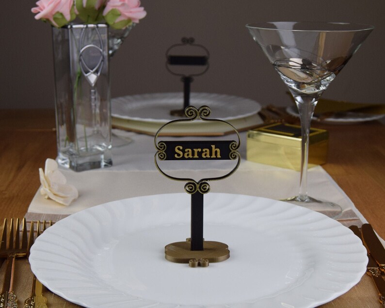 May include: A black and gold table place card with the name "Sarah" written in gold lettering. The place card is on a white plate with a gold fork and knife next to it.