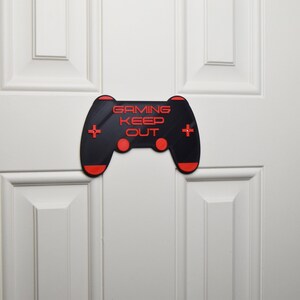 以下が含まれることがあります： 「Gaming Keep Out」というテキストが書かれた、ビデオゲームコントローラーの形をした黒と赤の3Dプリントされたドアサイン。