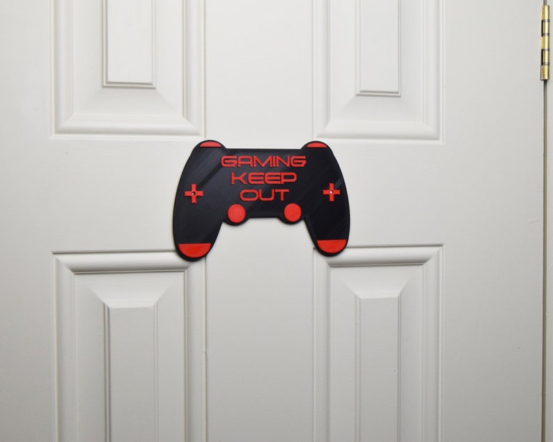 以下が含まれることがあります： 「Gaming Keep Out」というテキストが書かれた、ビデオゲームコントローラーの形をした黒と赤の3Dプリントされたドアサイン。