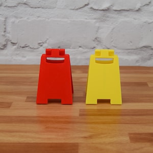 May include: Two caution signs, one red and one yellow, with a smiling face design. The signs are made of plastic and are standing on a wooden surface.