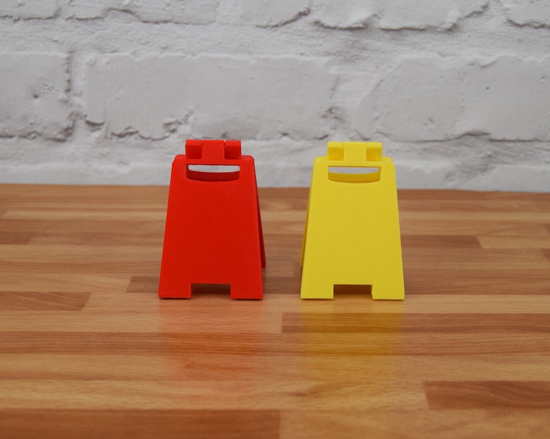 May include: Two caution signs, one red and one yellow, with a smiling face design. The signs are made of plastic and are standing on a wooden surface.