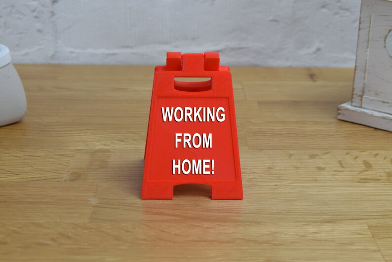 May include: A red, plastic, triangular sign with the text "WORKING FROM HOME!" in white letters.