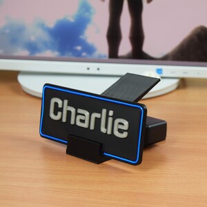 Pu&ograve; includere: Un'etichetta per scrivania nera con un contorno al neon blu e la parola "Charlie" in lettere argentate.