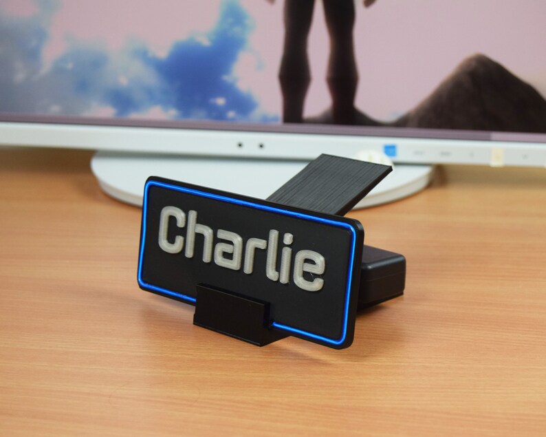Pu&ograve; includere: Un'etichetta per scrivania nera con un contorno al neon blu e la parola "Charlie" in lettere argentate.