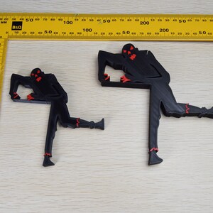 May include: Two black 3D printed figures of a zombie with red eyes and teeth. The figures are posed in a dynamic stance with one leg raised. The figures are on a light brown wooden surface.
