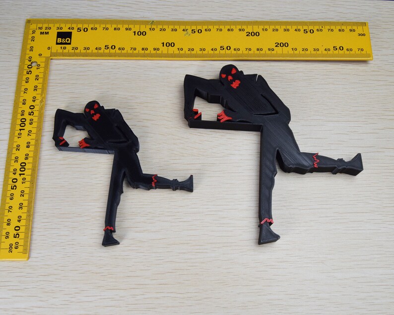 May include: Two black 3D printed figures of a zombie with red eyes and teeth. The figures are posed in a dynamic stance with one leg raised. The figures are on a light brown wooden surface.