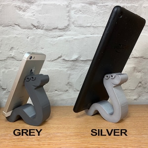 May include: Two phone stands made of 3D printed plastic in the shape of a cartoon worm. The stand on the left is gray and holds a smartphone. The stand on the right is silver and holds a tablet.