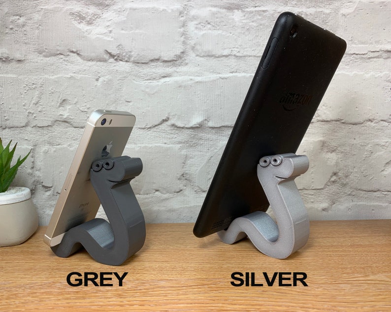 May include: Two phone stands made of 3D printed plastic in the shape of a cartoon worm. The stand on the left is gray and holds a smartphone. The stand on the right is silver and holds a tablet.