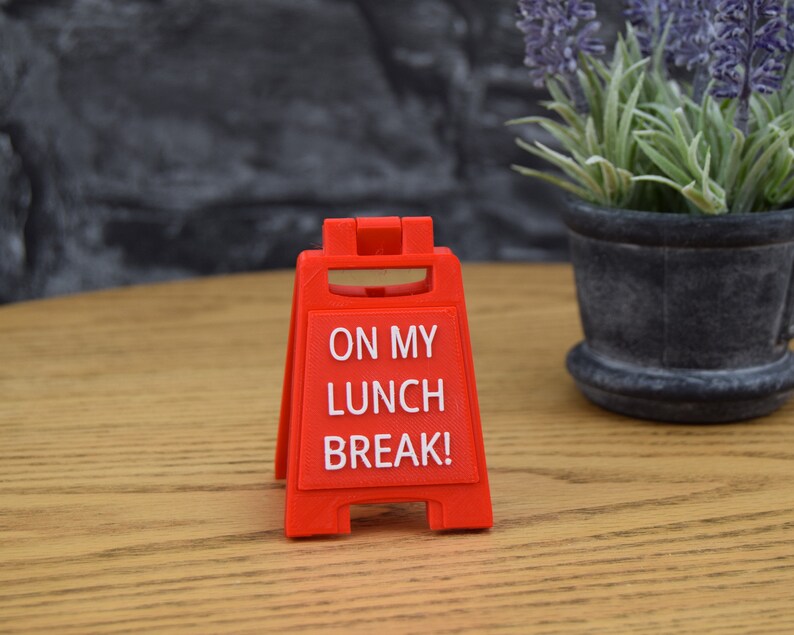 May include: A bright red, plastic sign with the text "ON MY LUNCH BREAK!" in white. The sign is in an A-frame style, designed to stand upright. The background includes a wooden table and a potted plant.