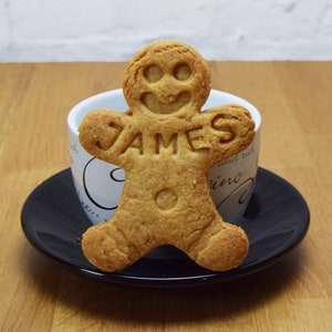 Puede incluir: Una galleta de jengibre con forma de persona con el nombre "JAMES" escrito en ella. La galleta est&aacute; sobre un platillo blanco con borde negro y un platillo negro.