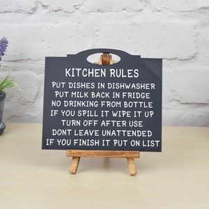 May include: Black chalkboard sign with white text on a wooden easel. The sign reads "Kitchen Rules" and lists rules for kitchen etiquette, such as "Put dishes in dishwasher" and "Turn off after use".