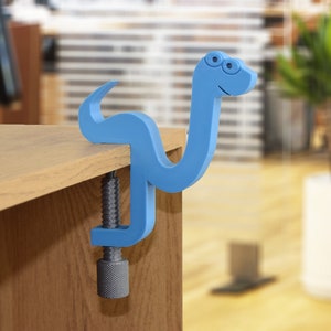 May include: A blue plastic clamp shaped like a snake with a smiling face. The clamp is attached to a wooden surface.