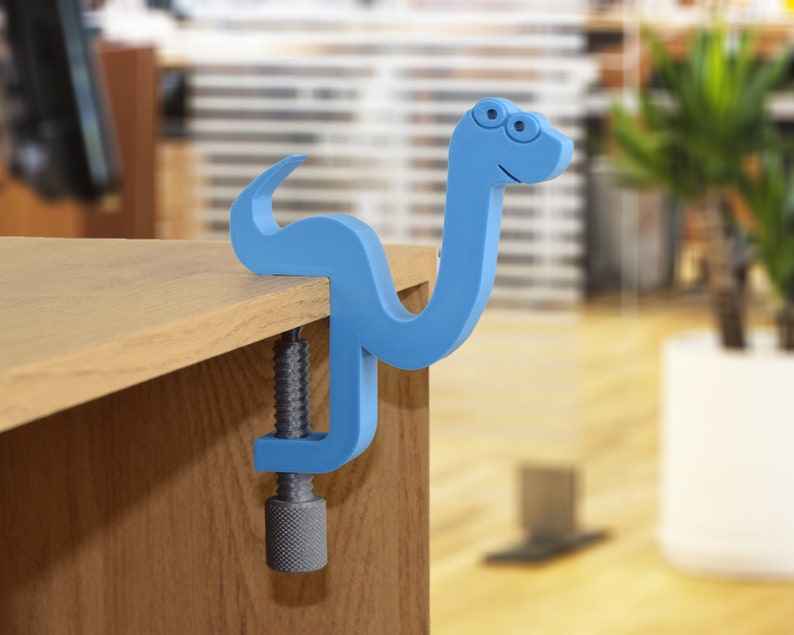 May include: A blue plastic clamp shaped like a snake with a smiling face. The clamp is attached to a wooden surface.