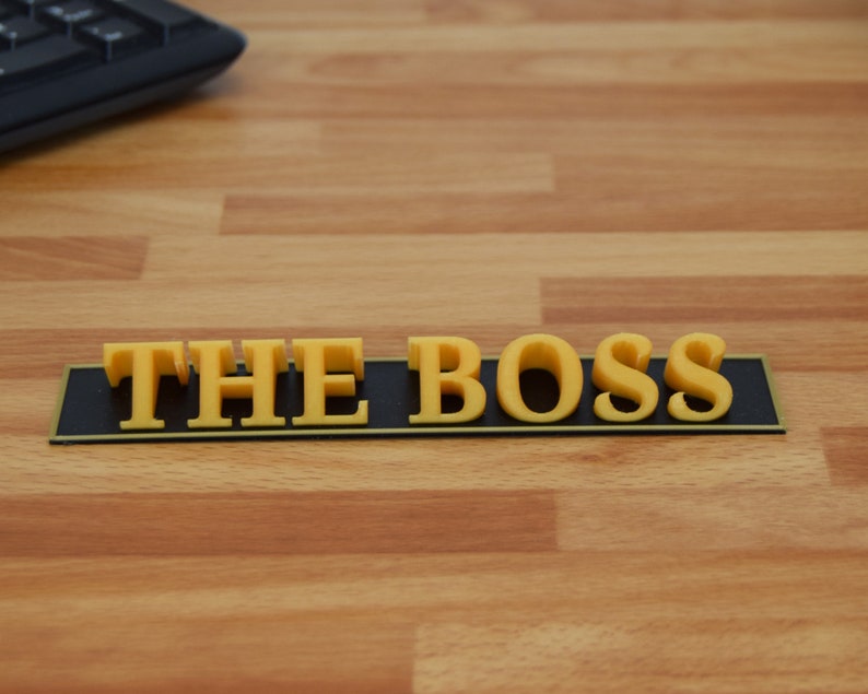 May include: A desk sign that reads "THE BOSS" in yellow 3D lettering. The letters are mounted on a black rectangular base with a thin gold border. The sign is on a light brown wooden desk.