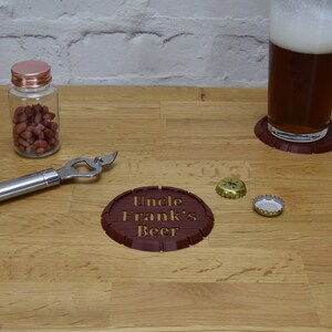 May include: A wooden table setting featuring a glass of beer on a coaster, a bottle of peanuts, a bottle opener, and a coaster with "Uncle Frank's Beer" text. Two bottle caps are also present. The coaster is approximately 4 inches in diameter.