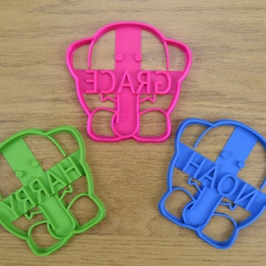 May include: Three cookie cutters shaped like elephants with the names "GRACE", "HARRY", and "HAOLI" written on them. The cutters are pink, green, and blue.