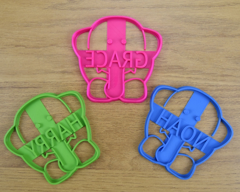 May include: Three cookie cutters shaped like elephants with the names "GRACE", "HARRY", and "HAOLI" written on them. The cutters are pink, green, and blue.