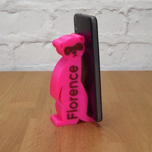 May include: A pink 3D printed phone stand shaped like a raccoon with the name "Florence" printed on the side. The stand is holding a black smartphone.