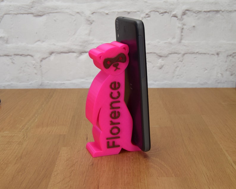 May include: A pink 3D printed phone stand shaped like a raccoon with the name "Florence" printed on the side. The stand is holding a black smartphone.
