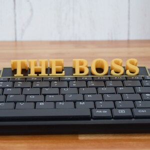 May include: A black computer keyboard with yellow 3D printed letters spelling "THE BOSS" across the top. The keyboard is on a wooden surface with a white background.