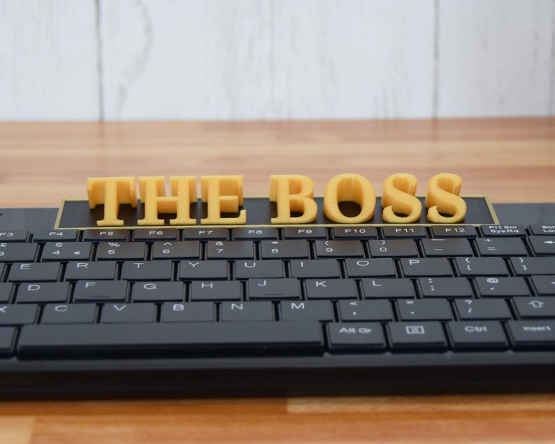 May include: A black computer keyboard with yellow 3D printed letters spelling "THE BOSS" across the top. The keyboard is on a wooden surface with a white background.