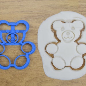 May include: Blue plastic cookie cutter shaped like a teddy bear. The cutter is on a wooden surface next to a white cookie dough cutout of a teddy bear.