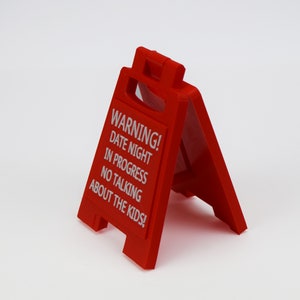 May include: A bright red, plastic A-frame sign with white text. The sign reads "WARNING! DATE NIGHT IN PROGRESS NO TALKING ABOUT THE KIDS!" The sign is designed to stand on its own, with a handle at the top.