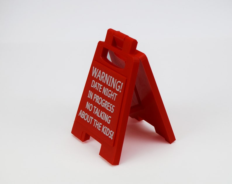 May include: A bright red, plastic A-frame sign with white text. The sign reads "WARNING! DATE NIGHT IN PROGRESS NO TALKING ABOUT THE KIDS!" The sign is designed to stand on its own, with a handle at the top.