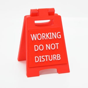 Op de afbeelding: Een rood plastic bord met witte tekst dat "WORKING DO NOT DISTURB" zegt.