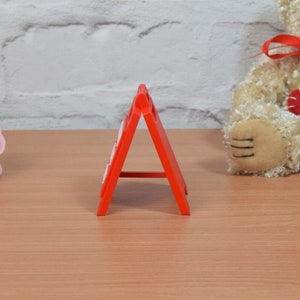 Op de afbeelding: Een rood, plastic A-frame bord met witte letters. Het bord is gevouwen en staat op een houten oppervlak.