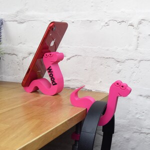 May include: Two pink worm-shaped phone and headphone holders. The phone holder has a red phone propped against it and the word "Wood" printed on the side.