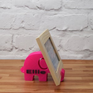 May include: A pink elephant-shaped photo stand with the name "Ellie" printed on it. A wooden picture frame is propped up against the stand.
