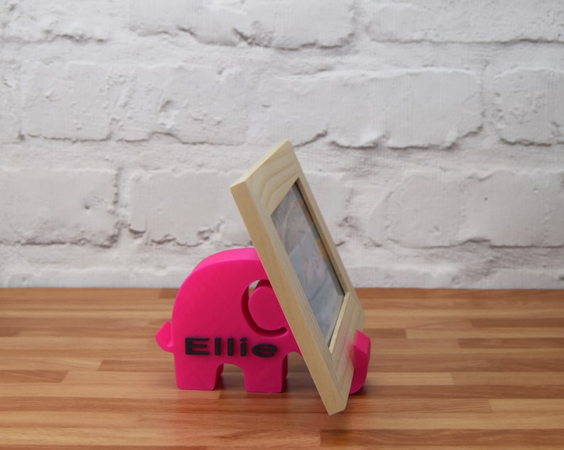 May include: A pink elephant-shaped photo stand with the name "Ellie" printed on it. A wooden picture frame is propped up against the stand.