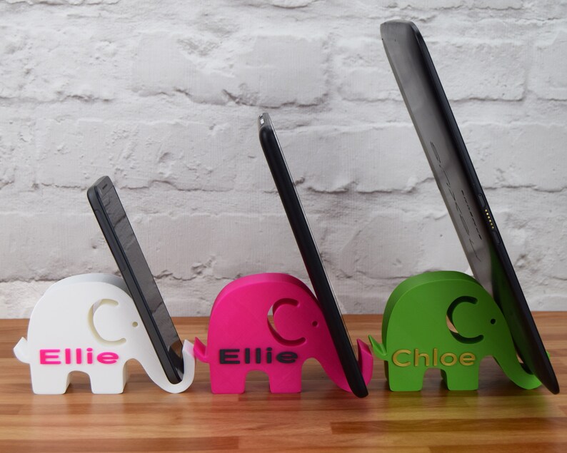 Pode incluir: Tr&ecirc;s suportes para telefone e tablet de elefante impressos em 3D. Os suportes s&atilde;o brancos, rosa e verdes. O suporte branco tem o nome "Ellie C." impresso nele. O suporte rosa tem o nome "Ellie C." impresso nele. O suporte verde tem o nome "Chloe C." impresso nele.