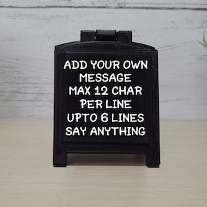 May include: A small, black chalkboard sign with a hinged top. The sign displays white text that reads "ADD YOUR OWN MESSAGE MAX 12 CHAR PER LINE UPTO 6 LINES SAY ANYTHING."