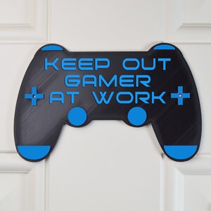 以下が含まれることがあります： 「Keep Out Gamer At Work」というテキストが書かれた、ビデオゲームコントローラーの形をした黒と青のゲーマー用ドアサイン。