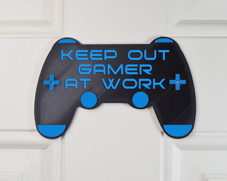 以下が含まれることがあります： 「Keep Out Gamer At Work」というテキストが書かれた、ビデオゲームコントローラーの形をした黒と青のゲーマー用ドアサイン。
