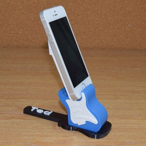 Puede incluir: Un soporte para tel&eacute;fono de color azul y blanco con forma de guitarra con el nombre "Ted" impreso en la base. Un tel&eacute;fono inteligente blanco est&aacute; en el soporte.