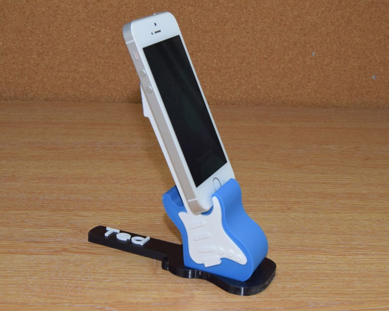 Puede incluir: Un soporte para tel&eacute;fono de color azul y blanco con forma de guitarra con el nombre "Ted" impreso en la base. Un tel&eacute;fono inteligente blanco est&aacute; en el soporte.