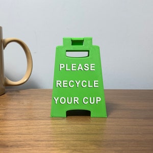 May include: A green plastic sign with the text "PLEASE RECYCLE YOUR CUP" in white letters.