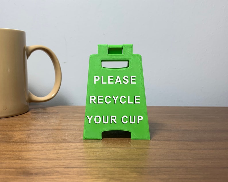 May include: A green plastic sign with the text "PLEASE RECYCLE YOUR CUP" in white letters.
