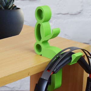 May include: A green 3D printed headphone stand shaped like a person sitting down. The stand has a hook on the back for hanging headphones. A pair of black headphones are hanging on the stand.