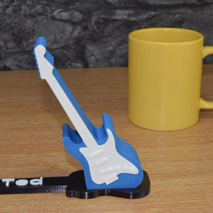 Puede incluir: Un soporte para tel&eacute;fono con forma de guitarra azul y blanco con el nombre "Ted" impreso en la base. El soporte est&aacute; sobre una superficie de madera junto a una taza amarilla.