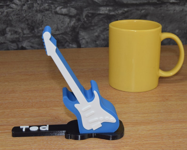 Puede incluir: Un soporte para tel&eacute;fono con forma de guitarra azul y blanco con el nombre "Ted" impreso en la base. El soporte est&aacute; sobre una superficie de madera junto a una taza amarilla.
