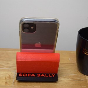 May include: A red phone stand with the words "SOFA SALLY" printed on the front. A clear phone case with a black phone inside is sitting in the stand.