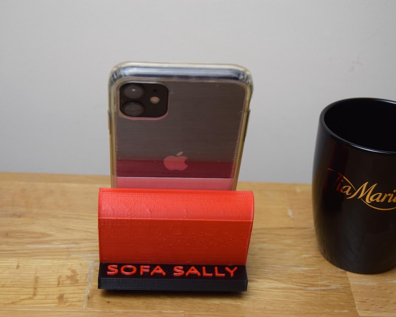 May include: A red phone stand with the words "SOFA SALLY" printed on the front. A clear phone case with a black phone inside is sitting in the stand.