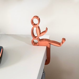 May include: A copper-colored 3D printed desk organizer shaped like a person sitting with their legs crossed and holding a phone. The organizer is mounted to a white desk.