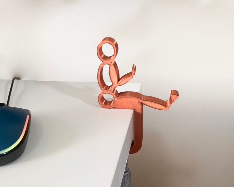 May include: A copper-colored 3D printed desk organizer shaped like a person sitting with their legs crossed and holding a phone. The organizer is mounted to a white desk.