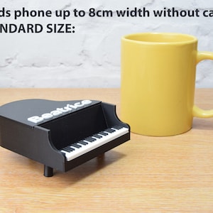 Puede incluir: Un soporte de tel&eacute;fono negro con forma de piano con la palabra "Beatrice" en la parte superior. El soporte est&aacute; al lado de una taza amarilla. El texto "Holds phone up to 8cm width without case STANDARD SIZE" es visible en la imagen.