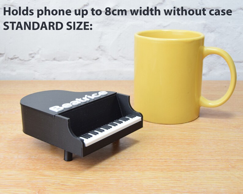 Puede incluir: Un soporte de tel&eacute;fono negro con forma de piano con la palabra "Beatrice" en la parte superior. El soporte est&aacute; al lado de una taza amarilla. El texto "Holds phone up to 8cm width without case STANDARD SIZE" es visible en la imagen.